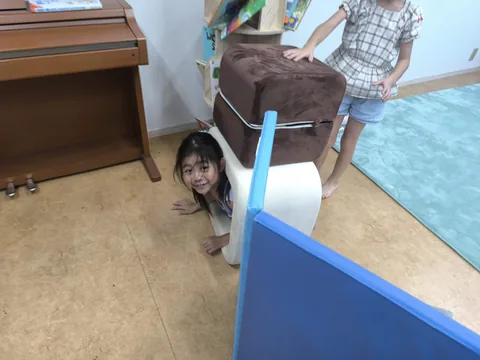 クッションの遊具の陰から笑顔をのぞかせるアフタースクールの子ども