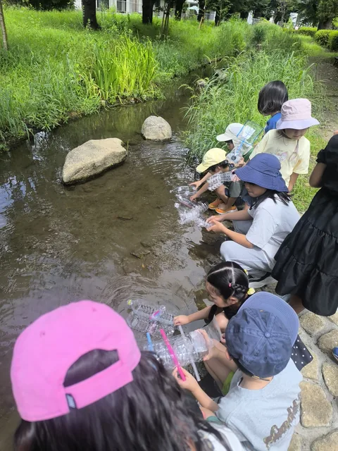 小川の水辺にしゃがんでペットボトルで実験をするアフタースクールの子どもたち