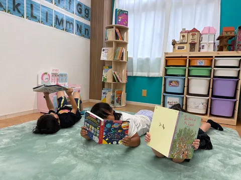 マットに寝転んで英語の絵本を読む子どもたち