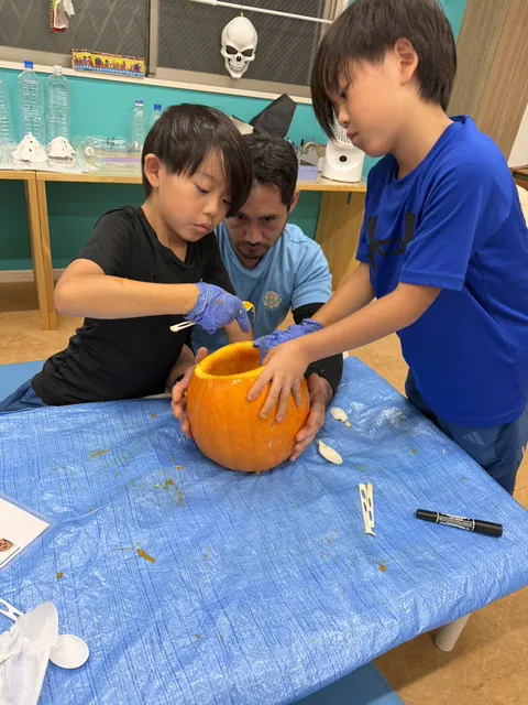 先生がかぼちゃを支え、スプーンで中身をきれいにくり抜く子どもたち