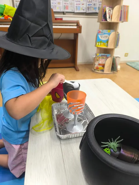 キンダースクールで魔女の帽子をかぶりハロウィンのカップでスプーンを使って遊ぶ子ども