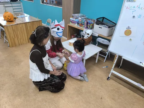 ピーカブーのハロウィンで、プリンセスのドレスを着た女の子がお母さんと先生と一緒にシールを選ぶ様子