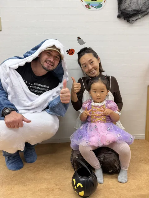 サメの着ぐるみの先生とお母さんと一緒に、ジャック・オー・ランタンのバケツを前に記念撮影する女の子