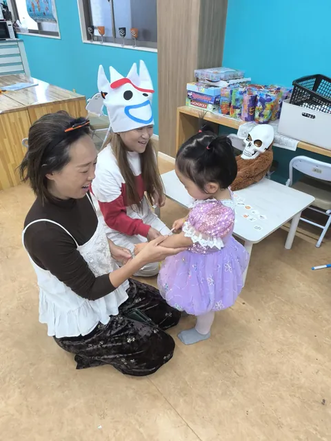 ハロウィンの仮装をした女の子の腕に、お母さんと先生がタトゥーシールを貼ってあげる様子