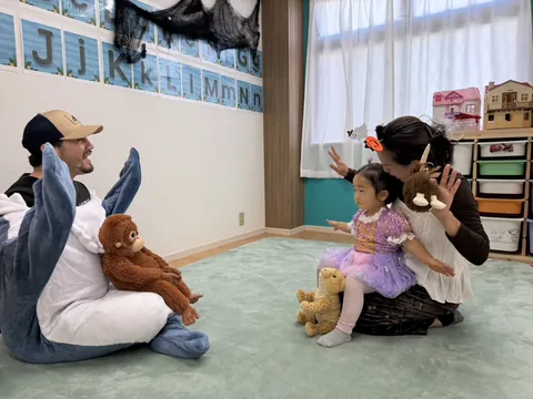 サメの着ぐるみの先生と向かい合い、お母さんのひざの上で手を広げて体を動かす女の子