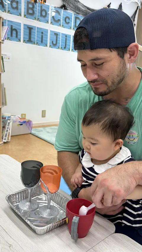 プリスクールで先生のひざの上でハロウィンのカップを使った色水あそびをする赤ちゃん
