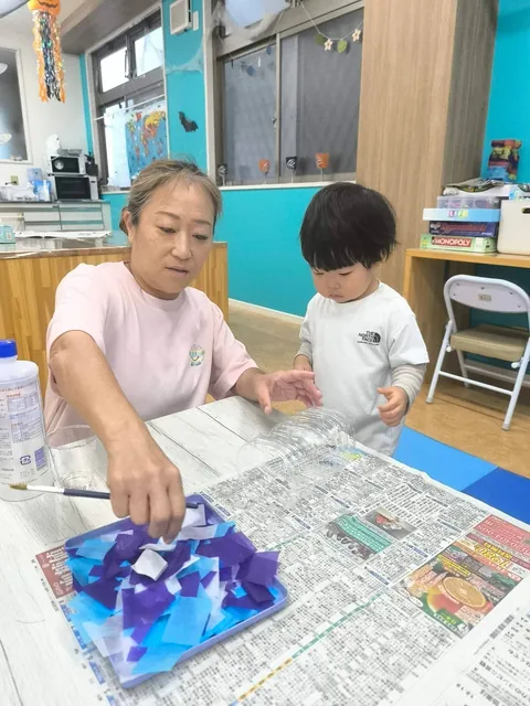 プリスクールで先生がのりを塗る様子を見つめる赤ちゃん