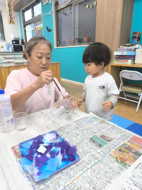 プリスクールで先生がペットボトルにのりを塗るのをじっと見る赤ちゃん