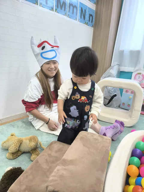 ハロウィンの帽子をかぶった先生のそばで、ハロウィン柄のオーバーオールを着てクッションの坂に向かう赤ちゃん