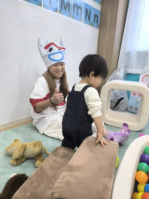 クッションの坂をのぼる赤ちゃんに、ハロウィンの帽子をかぶった先生がグッドサイン