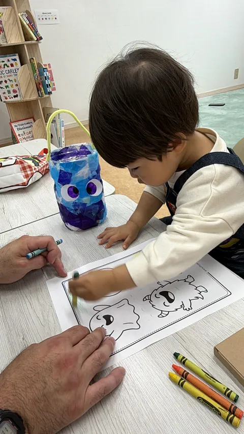 手作りのハロウィンバケツの横で、モンスターのぬり絵をする子ども