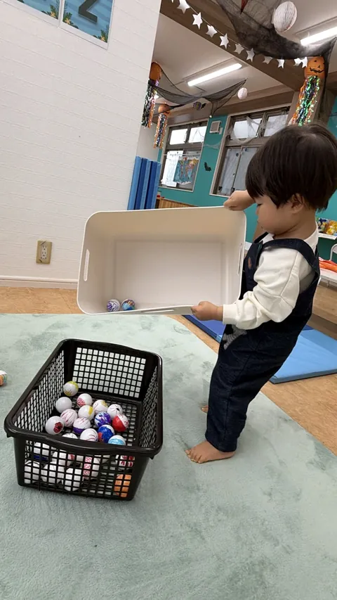 ボールの入ったかごの横で、箱を持ち上げてお片づけをする子ども