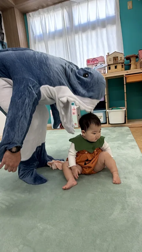 サメの着ぐるみの先生がのぞきこむ中、すわって遊ぶかぼちゃの衣装の赤ちゃん