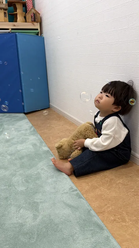 くまのぬいぐるみを抱っこしながら、ふわふわ飛ぶシャボン玉を見上げる子ども