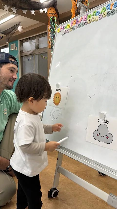 プリスクールでsunnyやcloudyのお天気カードの前でホワイトボードにお絵かきする赤ちゃん